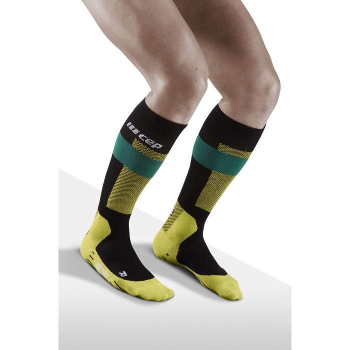 Lower legs of model crouching in knee-high CEP Merino skiing compression socks in green, yellow and black accents.